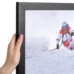 May include: A black picture frame containing a photograph of a skier in a white and red jacket, blue goggles, and a white helmet, carving through deep snow on a mountain slope, creating a spray of white powder as they move across the frame.