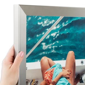 Puede incluir: Un marco rectangular de metal plateado con acabado cepillado, que muestra una fotograf&iacute;a de una persona con una camiseta azul claro y pantalones cortos estampados sentada en un barco, con agua turquesa y una barandilla blanca visible.