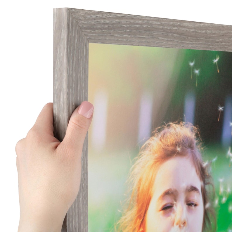 Puede incluir: Un marco de fotos rectangular con un acabado de grano de madera gris claro. El marco contiene una fotograf&iacute;a de una persona de cabello claro al aire libre, con tonos verdes y amarillos suaves de fondo y semillas de diente de le&oacute;n flotando.