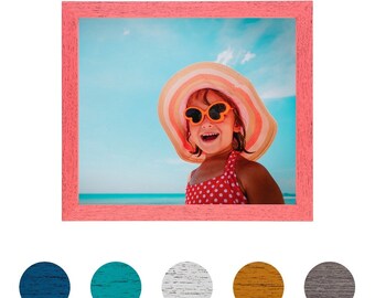 May include: A pink wood picture frame with a photo of a young girl wearing a colorful sun hat and sunglasses. The photo is taken outdoors with a blue sky and ocean in the background.