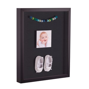 May include: A black shadow box frame with a white photo of a baby, a string of blue and green pom-poms, and a pair of white baby shoes.