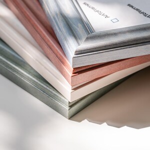 May include: A stack of picture frames in various colors, including gray, blue, pink, and white. The frames have a textured wood grain appearance. The text "Art of Frames" is visible on the top frame.