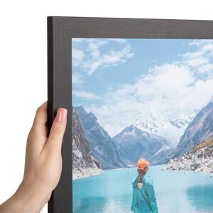 Puede incluir: Un marco de fotos de madera de color marr&oacute;n oscuro que contiene una fotograf&iacute;a de una persona con una gorra naranja y una chaqueta de color verde azulado, frente a un lago turquesa rodeado de monta&ntilde;as nevadas bajo un cielo azul.