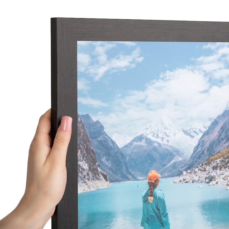 Puede incluir: Un marco de fotos de madera de color marr&oacute;n oscuro que contiene una fotograf&iacute;a de una persona con una gorra naranja y una chaqueta de color verde azulado, frente a un lago turquesa rodeado de monta&ntilde;as nevadas bajo un cielo azul.
