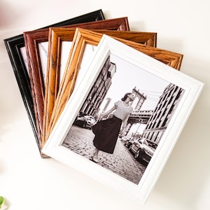 May include: A collection of rectangular picture frames in various colors, including black, brown, and white. The frames are made of wood and have a classic design. One frame displays a black and white photo of a woman in a city.