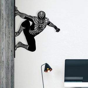 Op de afbeelding: Een zwarte metalen wanddecoratie van een superheldenfiguur in een klimpose. De decoratie is gemonteerd op een witte muur naast een houten muur. Een zwarte bureau met een zwarte stoel en een computermonitor staat op de voorgrond.