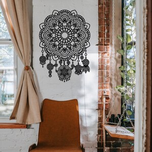May include: A black metal mandala wall decor with intricate details and dangling charms. The mandala is hanging on a white brick wall with a brown chair in the foreground.