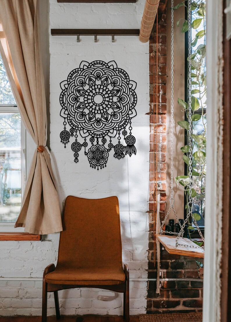 May include: A black metal mandala wall decor with intricate details and dangling charms. The mandala is hanging on a white brick wall with a brown chair in the foreground.