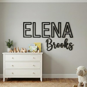 Puede incluir: Letrero de nombre de madera negra que dice "ELENA Brooks" en un entorno de guarder&iacute;a. El nombre est&aacute; en una fuente en negrita y en bloque, con el apellido en cursiva. Una c&oacute;moda blanca y una oveja de juguete est&aacute;n en primer plano.