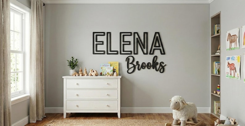 Puede incluir: Letrero de nombre de madera negra que dice "ELENA Brooks" en un entorno de guarder&iacute;a. El nombre est&aacute; en una fuente en negrita y en bloque, con el apellido en cursiva. Una c&oacute;moda blanca y una oveja de juguete est&aacute;n en primer plano.