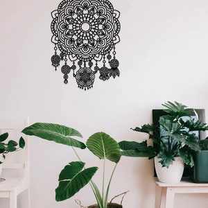 May include: Black metal wall decor featuring a large mandala design with intricate details and dangling charms. The mandala is surrounded by a white wall and a potted plant.