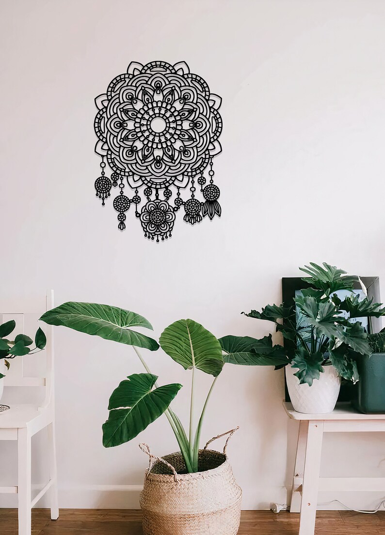 May include: Black metal wall decor featuring a large mandala design with intricate details and dangling charms. The mandala is surrounded by a white wall and a potted plant.