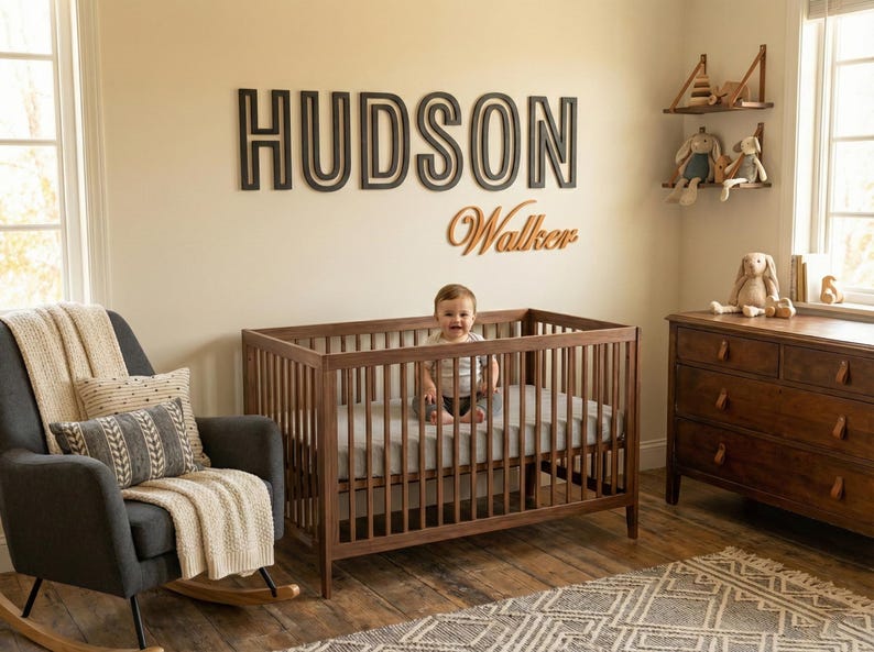 Puede incluir: Una escena de guarder&iacute;a con una cuna de madera, un sill&oacute;n mecedora gris oscuro y una c&oacute;moda de madera. El nombre "HUDSON" se muestra en la pared sobre la cuna, con "Walker" debajo. Un beb&eacute; est&aacute; en la cuna.