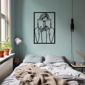 Puede incluir: Arte de pared de metal negro que representa a una persona leyendo un libro. El arte est&aacute; enmarcado en un cuadrado negro y est&aacute; montado en una pared azul claro.