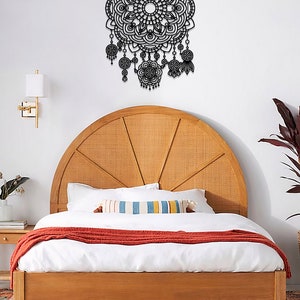 May include: A black metal mandala wall decor with intricate details and dangling beads. The decor is hanging on a white wall above a wooden bed with a white duvet and a red throw blanket.