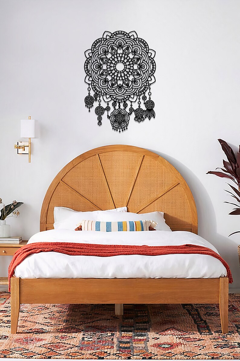 May include: A black metal mandala wall decor with intricate details and dangling beads. The decor is hanging on a white wall above a wooden bed with a white duvet and a red throw blanket.