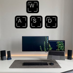 May include: A white desk with a black keyboard, a mouse, a laptop, and a monitor. There are two black speakers on the desk. On the wall behind the desk are four black square stickers with white letters that spell out "WASD".