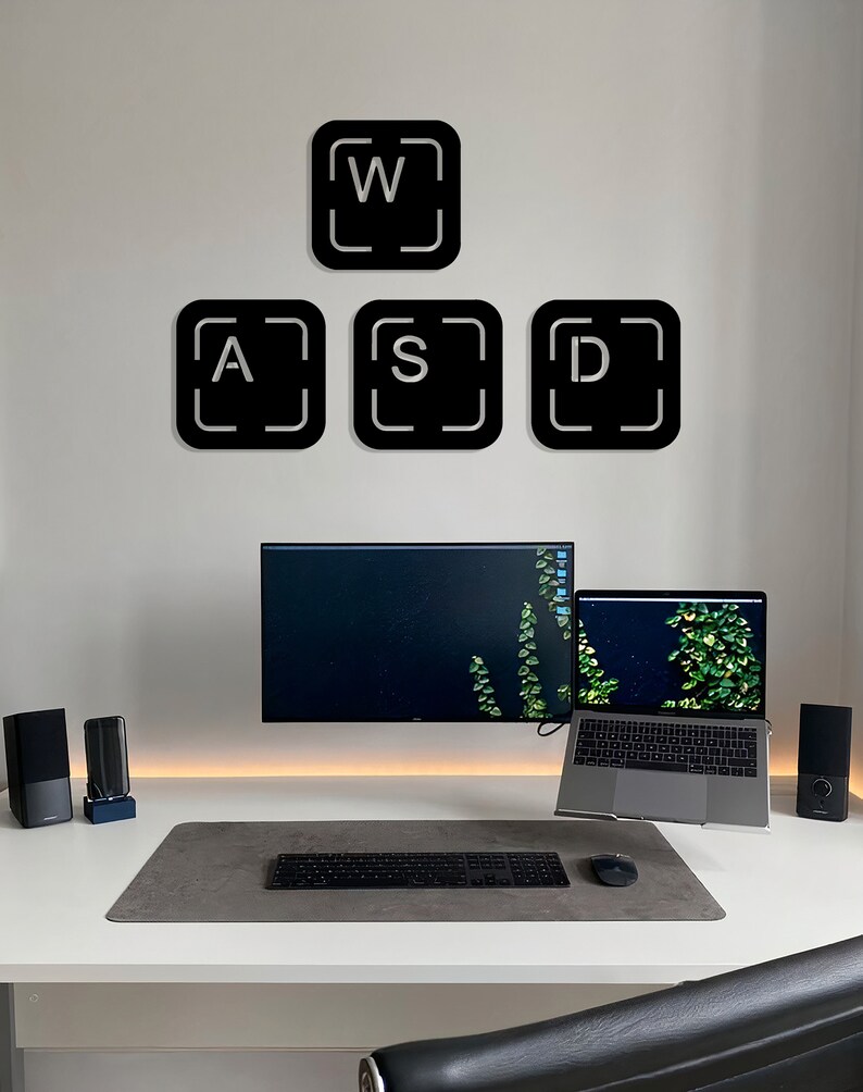 May include: A white desk with a black keyboard, a mouse, a laptop, and a monitor. There are two black speakers on the desk. On the wall behind the desk are four black square stickers with white letters that spell out "WASD".