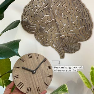 Puede incluir: Un reloj de madera con forma de cerebro con n&uacute;meros romanos. El reloj est&aacute; colgado en una pared y el texto "Puedes colgar el reloj donde quieras." est&aacute; escrito debajo.