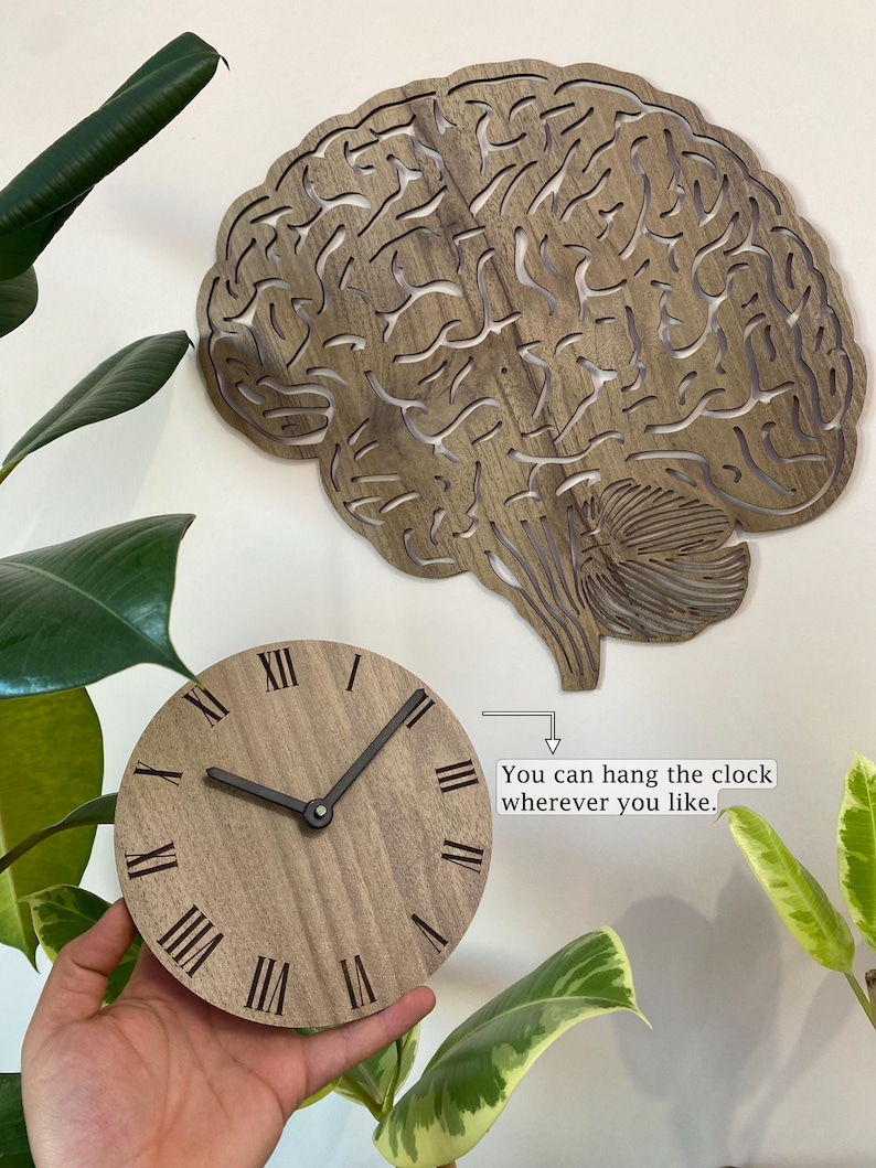 Puede incluir: Un reloj de madera con forma de cerebro con n&uacute;meros romanos. El reloj est&aacute; colgado en una pared y el texto "Puedes colgar el reloj donde quieras." est&aacute; escrito debajo.