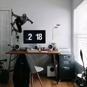 Op de afbeelding: Een zwarte metalen wandsculptuur van een superheldenfiguur, een bureau met een computermonitor, een kalender en een printer in een modern thuiskantoor.
