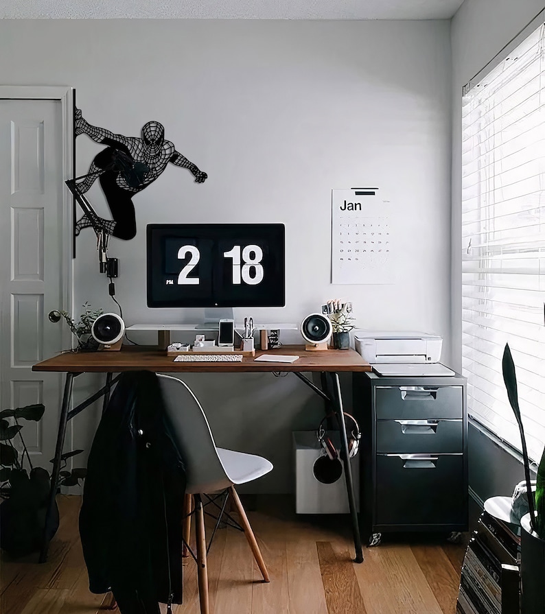 Op de afbeelding: Een zwarte metalen wandsculptuur van een superheldenfiguur, een bureau met een computermonitor, een kalender en een printer in een modern thuiskantoor.