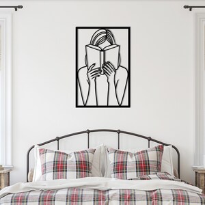 Puede incluir: Arte de pared de metal negro que representa a una persona leyendo un libro. El arte est&aacute; enmarcado y cuelga de una pared blanca sobre una cama con ropa de cama a cuadros rojos y blancos.