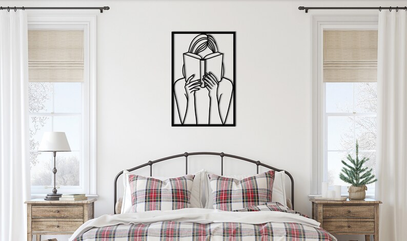 Puede incluir: Arte de pared de metal negro que representa a una persona leyendo un libro. El arte est&aacute; enmarcado y cuelga de una pared blanca sobre una cama con ropa de cama a cuadros rojos y blancos.