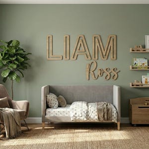 Puede incluir: Un cuarto de ni&ntilde;os con una cuna gris, estanter&iacute;as de madera y un sill&oacute;n mecedor. La pared muestra el nombre "LIAM Ross" en letras de madera. Una gran planta verde y una peque&ntilde;a mesa de madera completan la decoraci&oacute;n.