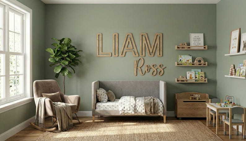 Puede incluir: Un cuarto de ni&ntilde;os con una cuna gris, estanter&iacute;as de madera y un sill&oacute;n mecedor. La pared muestra el nombre "LIAM Ross" en letras de madera. Una gran planta verde y una peque&ntilde;a mesa de madera completan la decoraci&oacute;n.