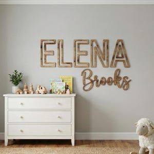 Puede incluir: Una habitaci&oacute;n infantil con una c&oacute;moda blanca, juguetes de madera y un oveja balanc&iacute;n. La pared presenta letras de madera que deletrean "ELENA Brooks". Una ventana con cortinas beige est&aacute; a la izquierda y una estanter&iacute;a a la derecha.