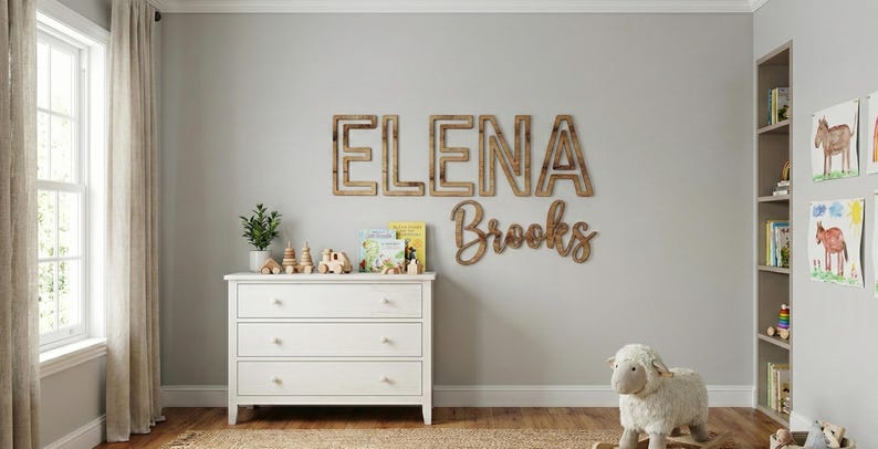 Puede incluir: Una habitaci&oacute;n infantil con una c&oacute;moda blanca, juguetes de madera y un oveja balanc&iacute;n. La pared presenta letras de madera que deletrean "ELENA Brooks". Una ventana con cortinas beige est&aacute; a la izquierda y una estanter&iacute;a a la derecha.