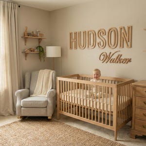 Puede incluir: Una habitaci&oacute;n infantil con una cuna de madera, una c&oacute;moda y estanter&iacute;as. El nombre "HUDSON Walker" se muestra en la pared. Un beb&eacute; est&aacute; sentado en la cuna. La habitaci&oacute;n tiene una paleta de colores neutros con una alfombra y una mecedora.