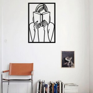 Puede incluir: Decoraci&oacute;n de pared de arte de l&iacute;nea en blanco y negro de una persona leyendo un libro. El arte est&aacute; enmarcado en un marco negro y est&aacute; colgado en una pared blanca.