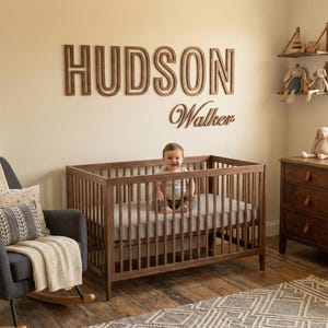 Puede incluir: Un cuarto de beb&eacute; con una cuna de madera, un sill&oacute;n mecedora gris oscuro y una c&oacute;moda de madera. La pared muestra el nombre "HUDSON Walker" en letras de madera. Un beb&eacute; est&aacute; sentado en la cuna.