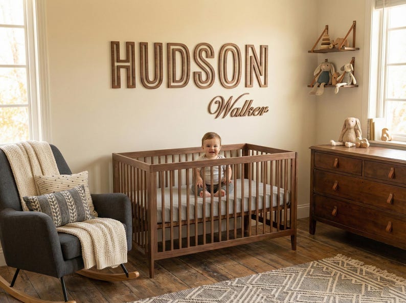 Puede incluir: Un cuarto de beb&eacute; con una cuna de madera, un sill&oacute;n mecedora gris oscuro y una c&oacute;moda de madera. La pared muestra el nombre "HUDSON Walker" en letras de madera. Un beb&eacute; est&aacute; sentado en la cuna.