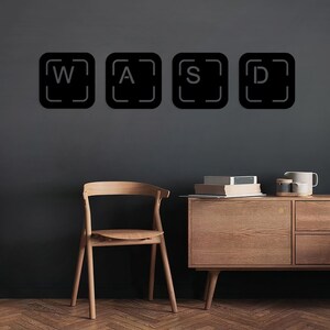 May include: Black and white keyboard keys with the letters W, A, S, and D on a grey wall. A wooden chair and a wooden cabinet with drawers are in the background.