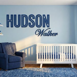 Puede incluir: Un cuarto de beb&eacute; con una tem&aacute;tica azul y blanca. La pared presenta el nombre "HUDSON" sobre el nombre "Walker" en una fuente cursiva. Una cuna blanca, un sill&oacute;n azul y una alfombra azul completan la habitaci&oacute;n.