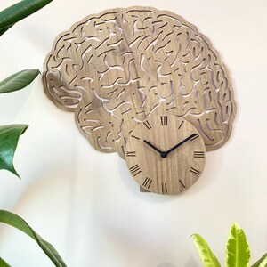 Puede incluir: Un reloj de pared de madera con forma de cerebro con n&uacute;meros romanos. La esfera del reloj est&aacute; en el centro de la forma del cerebro.
