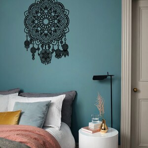 May include: A black metal mandala wall decoration with intricate details and dangling charms. The decor is hanging on a teal wall above a bed with a white duvet, grey and white pillows, and a gold throw blanket. A white bedside table with a vase of dried flowers and a glass of water sits next to the bed.