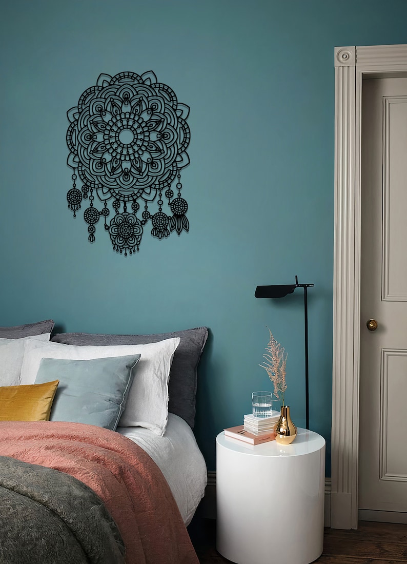 May include: A black metal mandala wall decoration with intricate details and dangling charms. The decor is hanging on a teal wall above a bed with a white duvet, grey and white pillows, and a gold throw blanket. A white bedside table with a vase of dried flowers and a glass of water sits next to the bed.