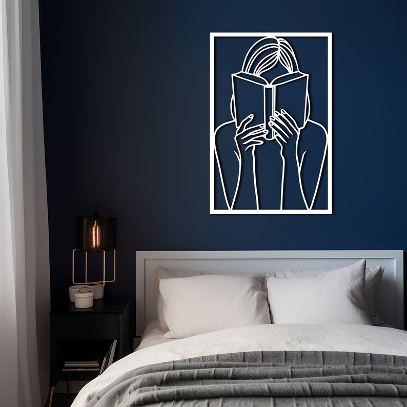 Puede incluir: Arte de pared de metal blanco que representa a una persona leyendo un libro. El rostro de la persona est&aacute; oculto por el libro. El arte est&aacute; enmarcado en un marco cuadrado blanco y est&aacute; montado en una pared azul oscuro.