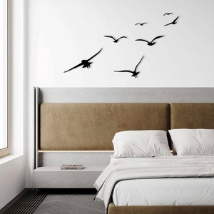 Puede incluir: Un interior de dormitorio con una cabecera marr&oacute;n y ropa de cama blanca. Una decoraci&oacute;n de pared de siluetas de p&aacute;jaros negros volando en bandada se muestra sobre la cama.