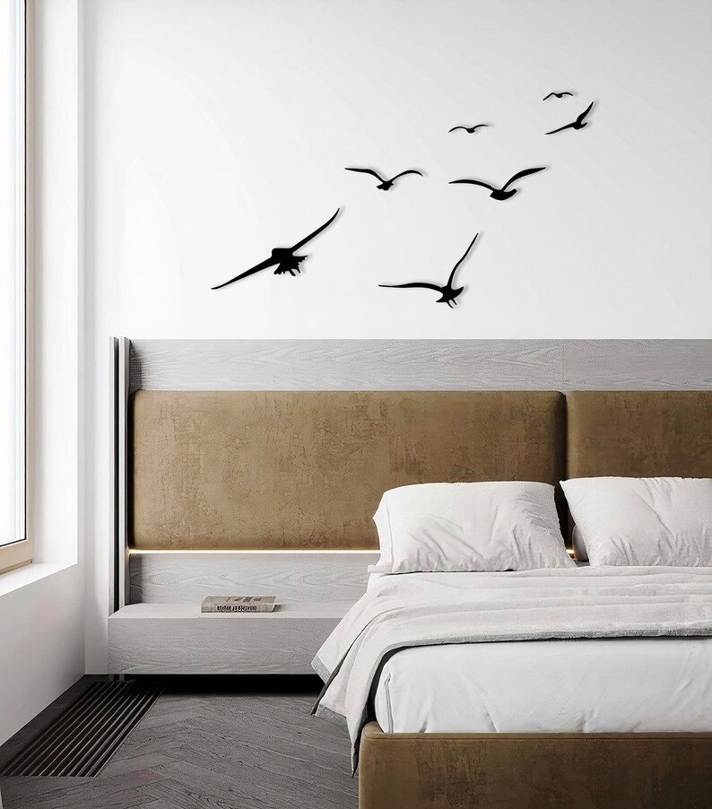 Puede incluir: Un interior de dormitorio con una cabecera marr&oacute;n y ropa de cama blanca. Una decoraci&oacute;n de pared de siluetas de p&aacute;jaros negros volando en bandada se muestra sobre la cama.