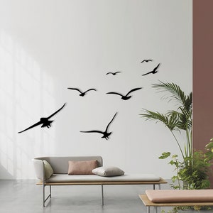 Puede incluir: Una pared blanca con una bandada de p&aacute;jaros negros en silueta volando en diferentes direcciones. Los p&aacute;jaros est&aacute;n dispuestos en un patr&oacute;n aleatorio, creando un dise&ntilde;o din&aacute;mico y art&iacute;stico. Un sof&aacute; marr&oacute;n claro con dos almohadas est&aacute; en primer plano.