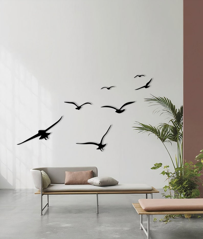 Puede incluir: Una pared blanca con una bandada de p&aacute;jaros negros en silueta volando en diferentes direcciones. Los p&aacute;jaros est&aacute;n dispuestos en un patr&oacute;n aleatorio, creando un dise&ntilde;o din&aacute;mico y art&iacute;stico. Un sof&aacute; marr&oacute;n claro con dos almohadas est&aacute; en primer plano.