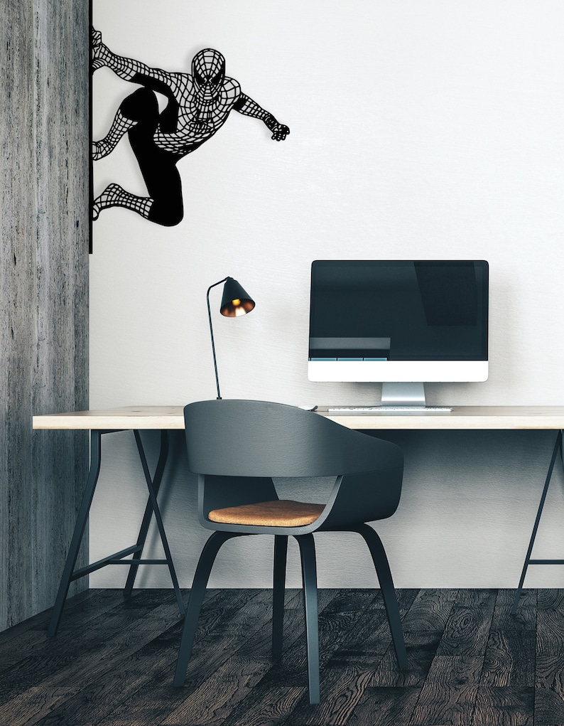 Op de afbeelding: Een zwarte metalen wanddecoratie van een superheldenfiguur in een klimpose. De decoratie is gemonteerd op een witte muur naast een houten muur. Een zwarte bureau met een zwarte stoel en een computermonitor staat op de voorgrond.