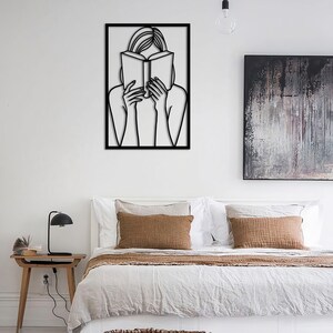 Puede incluir: Arte de pared de metal negro que representa a una persona leyendo un libro. El arte est&aacute; enmarcado en un marco cuadrado negro y est&aacute; colgado en una pared blanca.