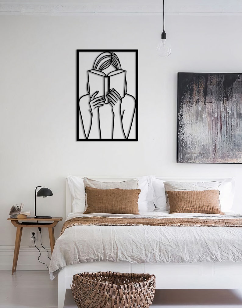 Puede incluir: Arte de pared de metal negro que representa a una persona leyendo un libro. El arte est&aacute; enmarcado en un marco cuadrado negro y est&aacute; colgado en una pared blanca.