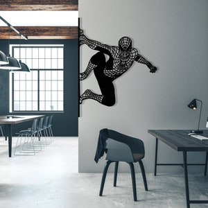 Op de afbeelding: Zwarte metalen wandkunst met een superheld in een web-pak, hangend aan een witte muur in een moderne kantooromgeving. De kunst is gemonteerd op een muur naast een bureau met een computer en een lamp.